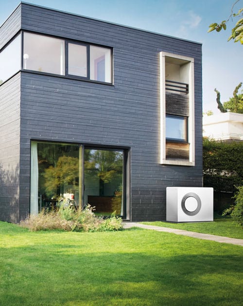 Modernes Einfamilienhaus mit großer Fensterfront und außen aufgestellter Wärmepumpe auf einer grünen Rasenfläche.